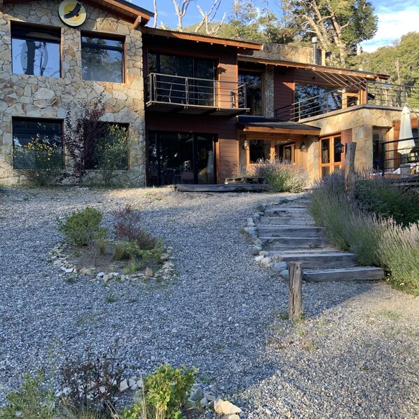 A two-story stone building with a gravel driveway and a wooden staircase leading to the entrance. - El Condor Pasa VLA (Villa La Angostura)