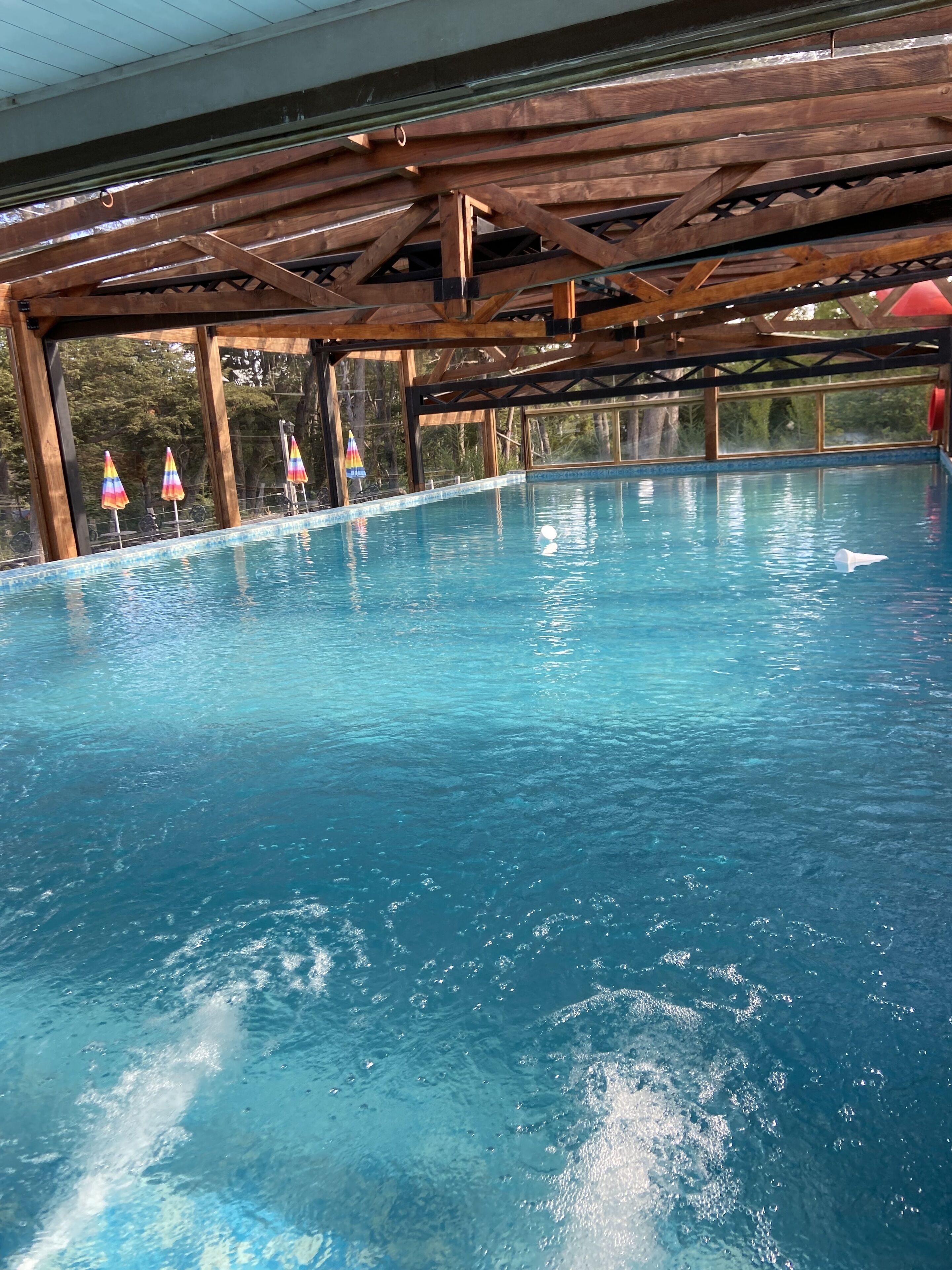 An indoor swimming pool with clear blue water, wooden beams overhead, and colorful umbrellas along the edge.