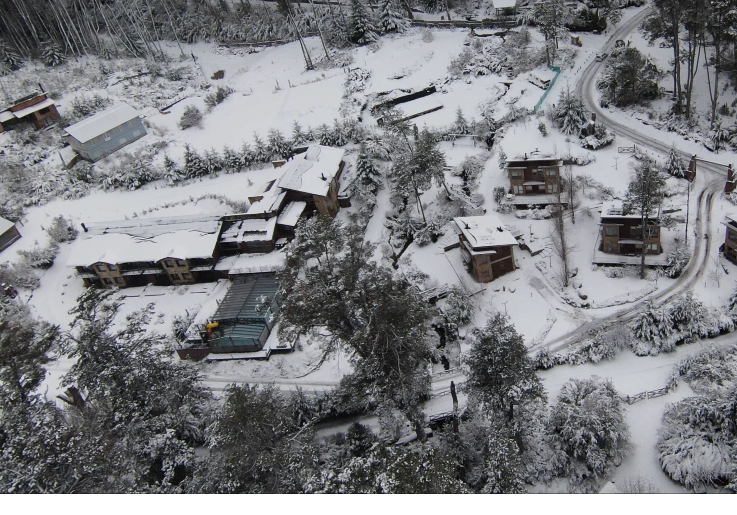 Un barrio residencial cubierto de nieve con varias casas, una carretera sinuosa y una pequeña zona de piscina.