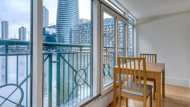 A room with a large window overlooking a river and cityscape, featuring a wooden table and chairs.
