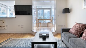 A modern living room with a grey sofa, a coffee table, and a TV mounted on the wall. There is a large window with a view of buildings and a balcony.