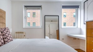 A bedroom with a large bed, two windows, a radiator, a wooden dresser, and a door leading to another room.