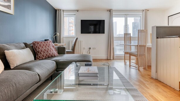 Un salon moderne avec un canapé gris, une table basse en verre et une vue sur la ville grâce à de grandes fenêtres.