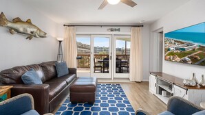 Un salon moderne avec un canapé en cuir, un pouf et un tapis à motifs bleus. On peut voir un ventilateur de plafond et une grande décoration représentant des poissons sur le mur.