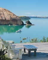 Una terraza de madera con sillones blancos y una mesa de picnic con vista al mar tranquilo, donde se aprecia una embarcación y una construcción con techo de paja.
