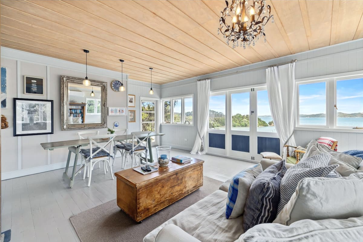 A spacious living room with a wooden ceiling, large windows with curtains, a dining area with white chairs, and a cozy seating area with a sofa and cushions.