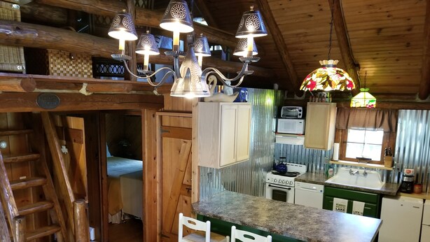 Una cocina rústica con armarios de madera, estufa y pileta. Hay tres sillas blancas y una escalera de madera.