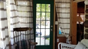 Una habitación acogedora con techo de madera, una puerta verde, cortinas a rayas, un sillón mecedor, un sofá blanco y una mesita pequeña.