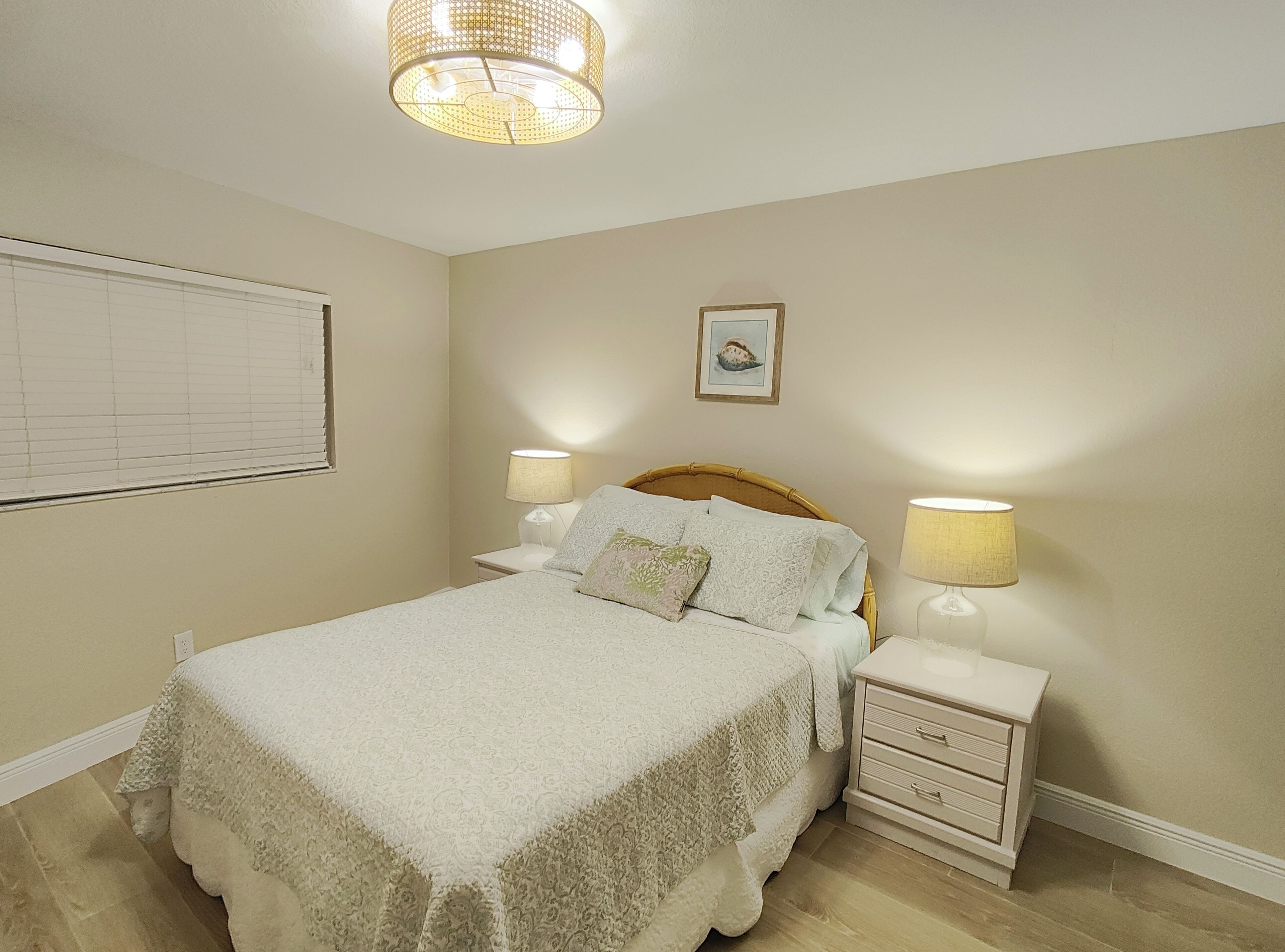 Un dormitorio con cama, dos mesitas de noche, lámpara, luz de techo, ventana con persianas y un cuadro en la pared.
