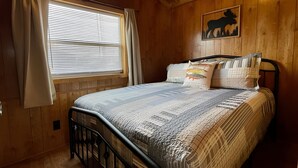 A wooden-paneled bedroom with a bed, curtains, a window with blinds, and a wall-mounted picture. - Cozy cabin in delightful Kemmerer with AC, WiFi (Kemmerer)