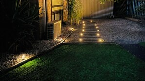 A well-lit residential property at dusk with a paved driveway leading to a garage, surrounded by greenery and a small lawn. - Stylish tiny home in Melton west (Melton West)