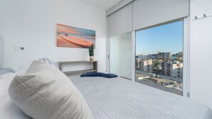 A modern hotel room with a large window, a bed, a bedside table, a painting, and a view of the city. - Elegant Penthouse with Private Terrace, BBQ & Jacuzzi (Cabo San Lucas)