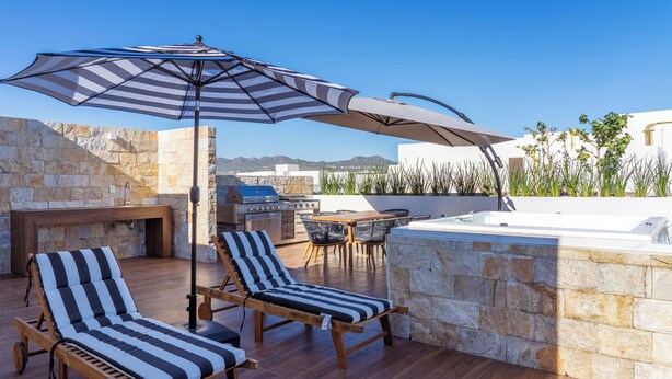 A rooftop terrace with two striped lounge chairs, a large umbrella, a stone wall, a barbecue area, and a view of the mountains. - Elegant Penthouse with Private Terrace, BBQ & Jacuzzi (Cabo San Lucas)