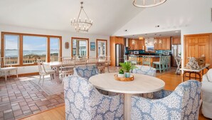 A spacious living area with a large round table, blue patterned chairs, a chandelier, and a view of the ocean through large windows. - 9 BR OCEAN/BEACHFRONT HOME w/ HEATEDPOOL & 2 HOTTUBS IN OBX! (Hatteras)
