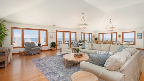 A spacious living room with a large sofa, a coffee table, and a view of the sea through large windows. - 9 BR OCEAN/BEACHFRONT HOME w/ HEATEDPOOL & 2 HOTTUBS IN OBX! (Hatteras)