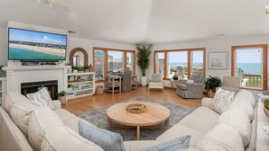 A spacious living room with a large TV, comfortable seating, a fireplace, and a view of the ocean. - 9 BR OCEAN/BEACHFRONT HOME w/ HEATEDPOOL & 2 HOTTUBS IN OBX! (Hatteras)