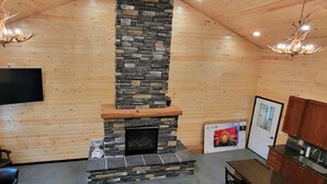 A log cabin interior with a stone fireplace, wooden walls, a mounted TV, and a kitchen area.