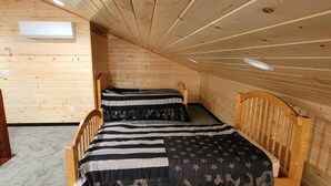 A small, cozy cabin room with two wooden beds, a sloped ceiling, and a built-in air conditioning unit.