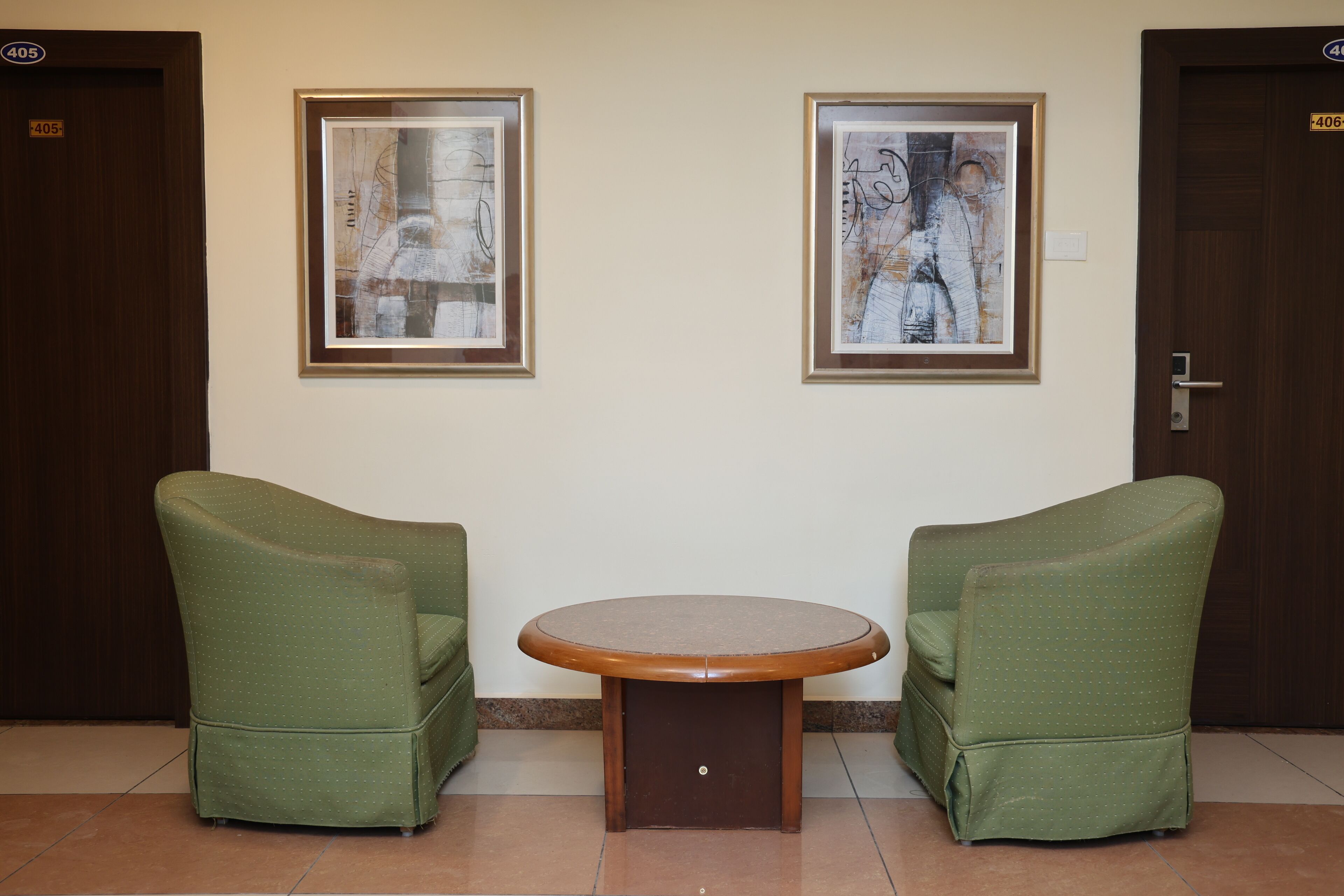 A room with two green armchairs, a small round table, and two framed abstract paintings on the wall.