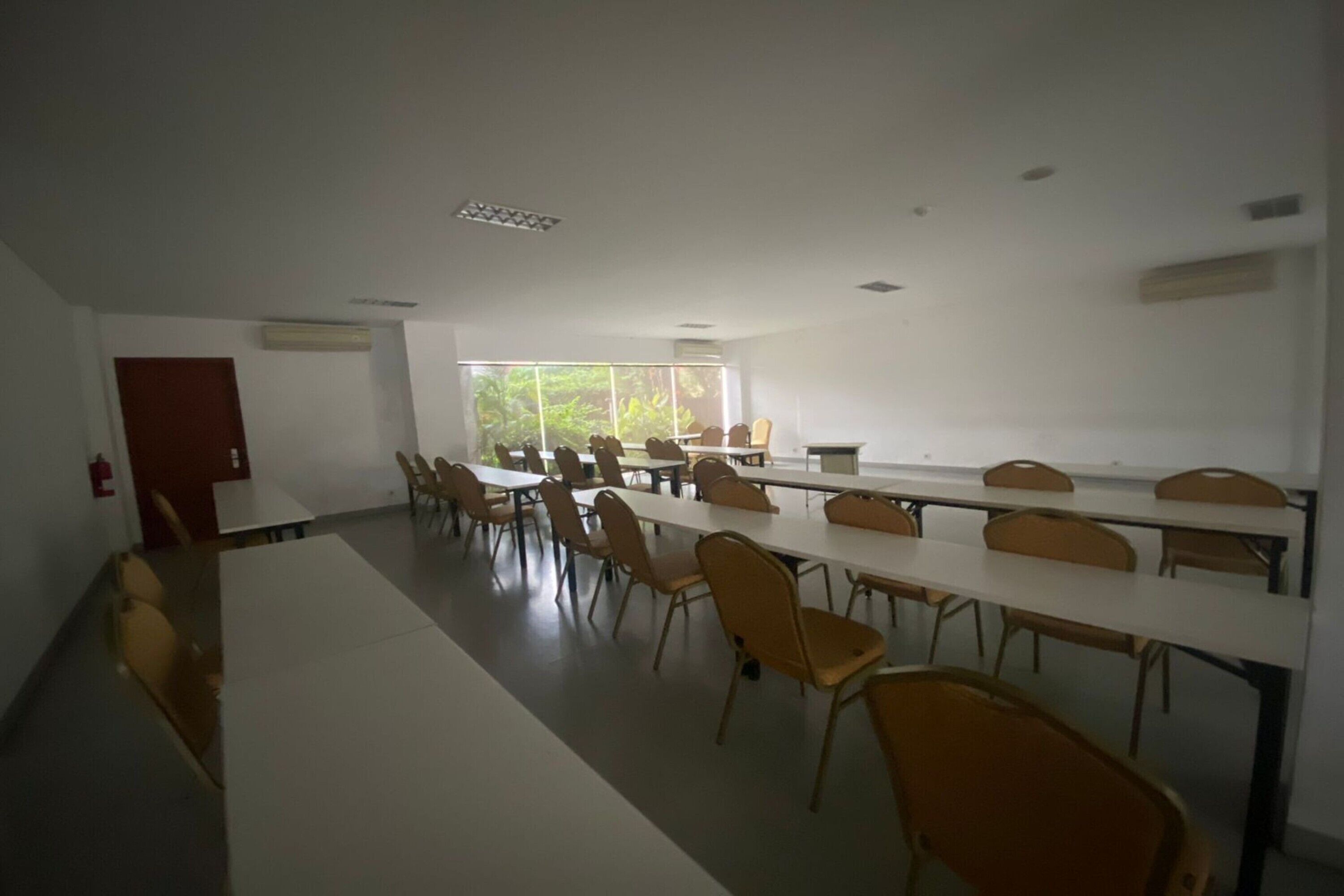 A conference room with long tables and chairs, large windows with a view of greenery, and a fire extinguisher on the wall.