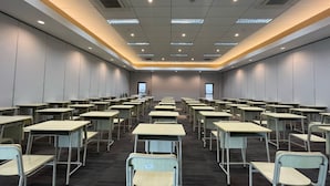 Une salle de conférence avec des rangées de tables et de chaises, un plafond doté d’un éclairage encastré et un mur sobre agrémenté d’une fenêtre.