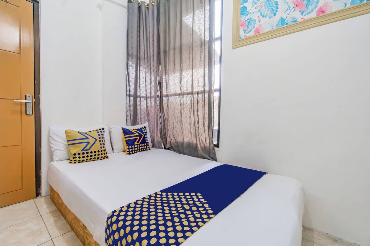 A neatly made bed with a blue and yellow patterned bedspread, two pillows with geometric designs, and a window with sheer curtains.