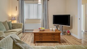 A living room with a sofa, a wooden coffee table with a bowl of fruit, a television, and a radiator. - Y Deri - Close to Village and Beach (Saundersfoot)