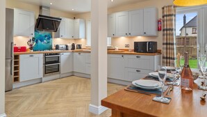 A modern kitchen with white cabinets, stainless steel appliances, and a wooden dining table set for six. - Y Deri - Close to Village and Beach (Saundersfoot)