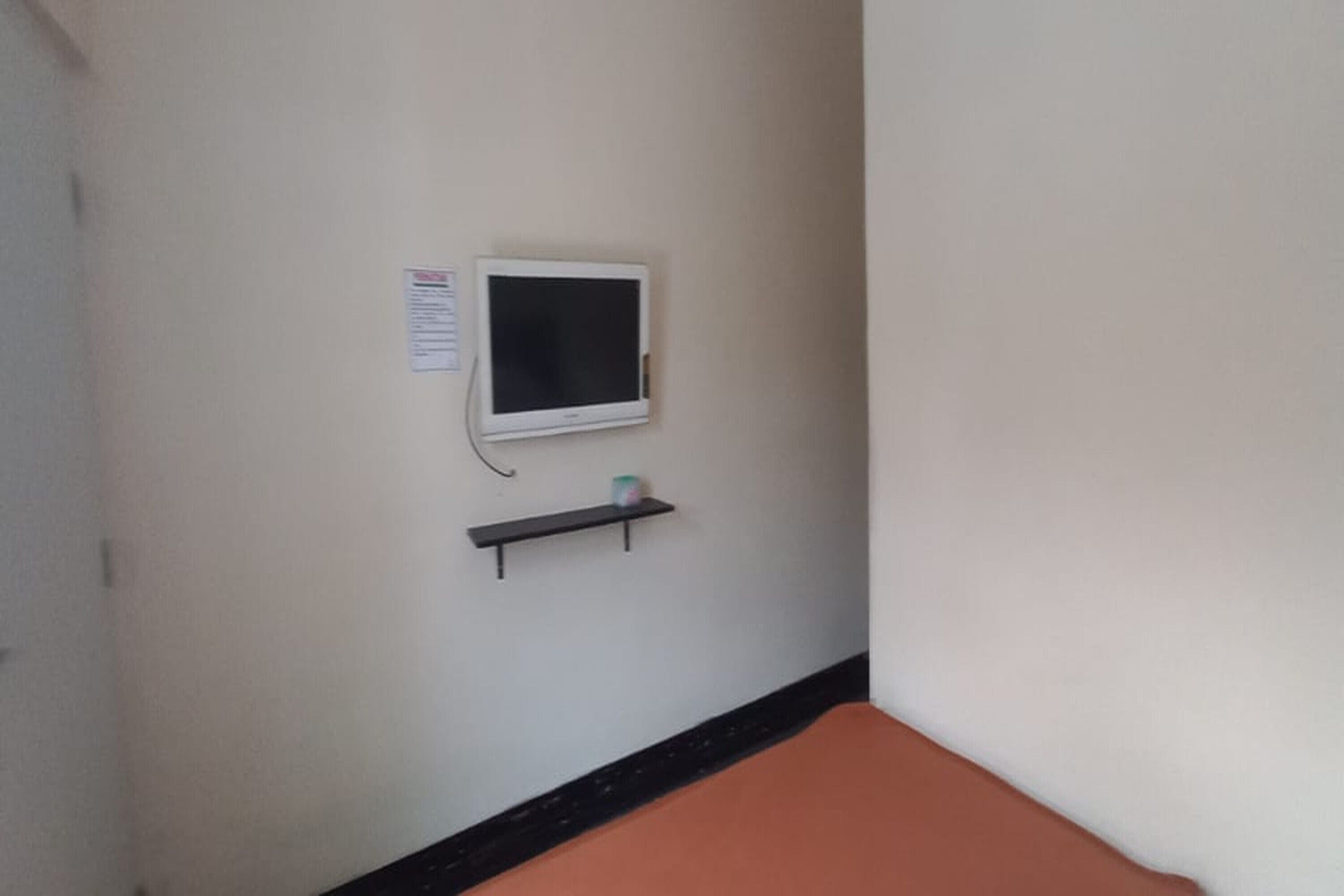 A room with a white wall, a small white TV mounted on the wall, and a black shelf below it.
