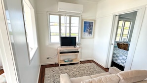 A room with a beige sofa, a TV on a stand, a window, and a door leading to another room.