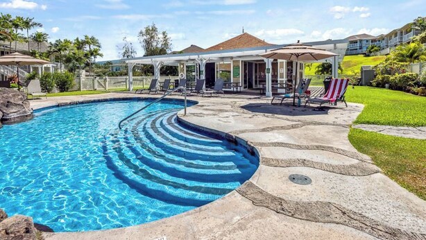 Une piscine à bord incurvé, un espace de détente couvert avec des chaises longues, et un bâtiment avec un toit en tuiles en arrière-plan.