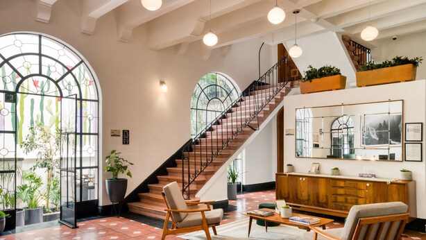A spacious lobby with a wooden staircase, large windows with plant decorations, and a wooden reception desk. - Santa Casa | Luxury King Room Suite in Roma Norte (Ciudad de México)