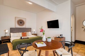 Une chambre d’hôtel comprenant un lit, une table de chevet avec une lampe, une table ronde en bois avec des chaises, un téléviseur fixé au mur et un vase à fleurs.