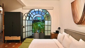 A hotel room with a bed, a nightstand, a lamp, a wardrobe, and a view through a window with a plant. - Santa Casa | Amazing King Room Central Roma Norte (Ciudad de México)