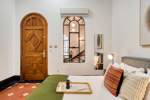 A room with a wooden door, a window with a view of a staircase, a bed with pillows, a tray with a book and mug, and a green rug. - Santa Casa | Cozy Queen Room in Central Roma Norte (Ciudad de México)