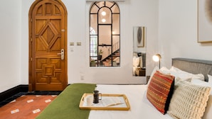 Ein Zimmer mit Holztür, einem Fenster mit Blick auf eine Treppe, einem Bett mit Kissen, einem Tablett mit Buch und Tasse sowie einem grünen Teppich.