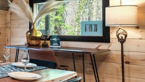 Et træhusinteriør med et spisebord dækket til to personer, en vase med fjer, en lampe og et vindue med udsigt til træer.