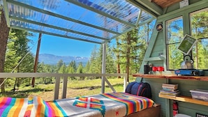 A cozy cabin with a bed, a desk with a lamp, a bookshelf, and a view of the forest and mountains. - A-Frame Glamping. Stargazing Near Missoula (Potomac)