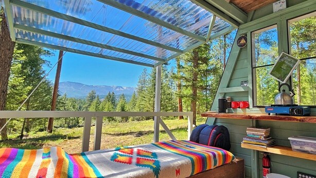 A cozy cabin with a bed, a desk with a lamp, a bookshelf, and a view of the forest and mountains. - A-Frame Glamping. Stargazing Near Missoula (Potomac)