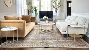 Ein Wohnzimmer mit einem beigefarbenen Sofa, einem weißen Sofa und einem runden Couchtisch. Im Hintergrund ist ein Fernseher zu sehen.