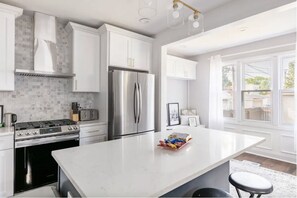 A modern kitchen with white cabinets, a stainless steel refrigerator, a gas stove, and a large island with a bowl of snacks. - Family friendly 5b/ 4ba sun-drenched home in Jefferson Park, 2 car garage! (Chicago)