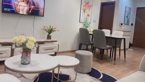 A modern living room with a flat-screen TV, a dining area with chairs and a table, and a vase with flowers on a round table.