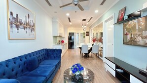 A modern living room with a blue sofa, a glass coffee table with blue flowers, a dining area with a white table and chairs, and a kitchen in the background.