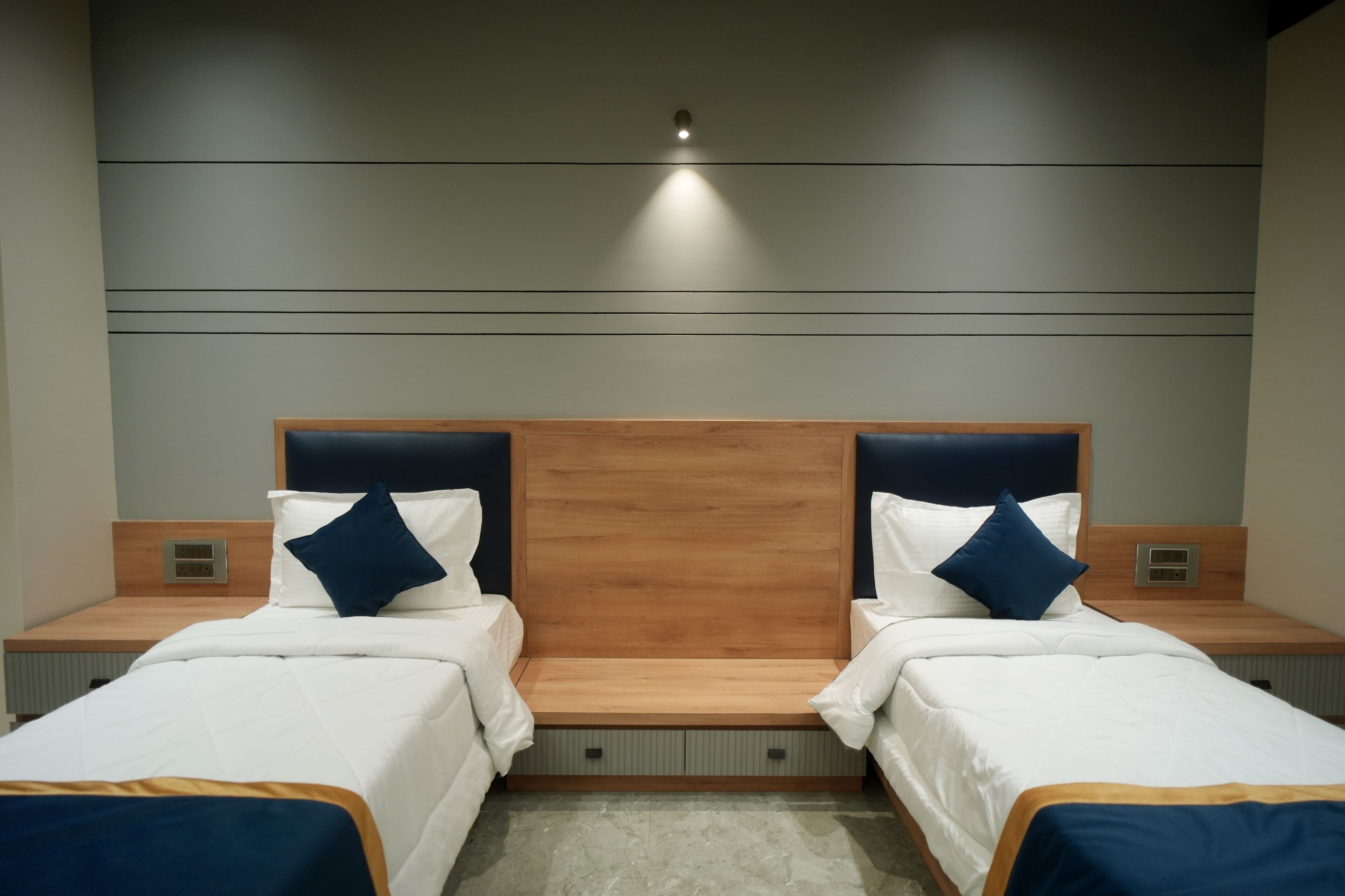 A hotel room with two beds, each with a wooden headboard, white bedding, and blue pillows. There are bedside tables with lamps and a small shelf.