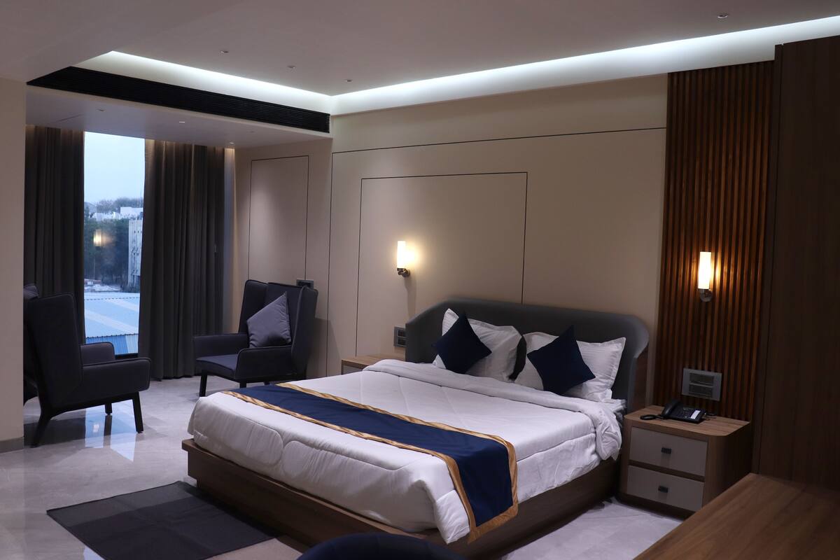 A modern hotel room with a large bed, a desk, and a chair. There is a window with curtains, a wall-mounted lamp, and a phone on the bedside table.