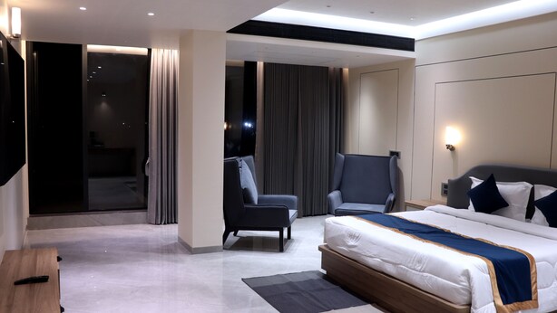 A modern hotel room with a large bed, two armchairs, a desk, and a sliding glass door leading to a balcony. - Dream Inn By Spring Tree (Wadhwan)
