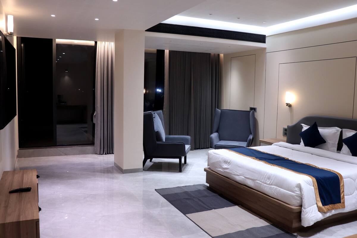 A modern hotel room with a large bed, two armchairs, a desk, and a sliding glass door leading to a balcony.