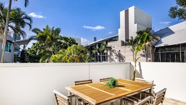 A rooftop terrace with a wooden table and chairs, surrounded by palm trees and modern buildings. - Central 2BR APT for 6PPL, w/ free parking@SouthBNE (South Brisbane)