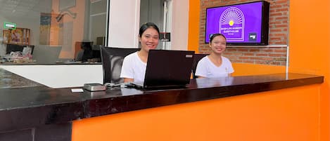 A reception desk with a laptop, a television displaying 'Babylon Angkor Guesthouse', and a brick wall.