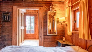 A wooden cabin interior with a bed, a mirror, a lamp, and a door leading to an outdoor area.